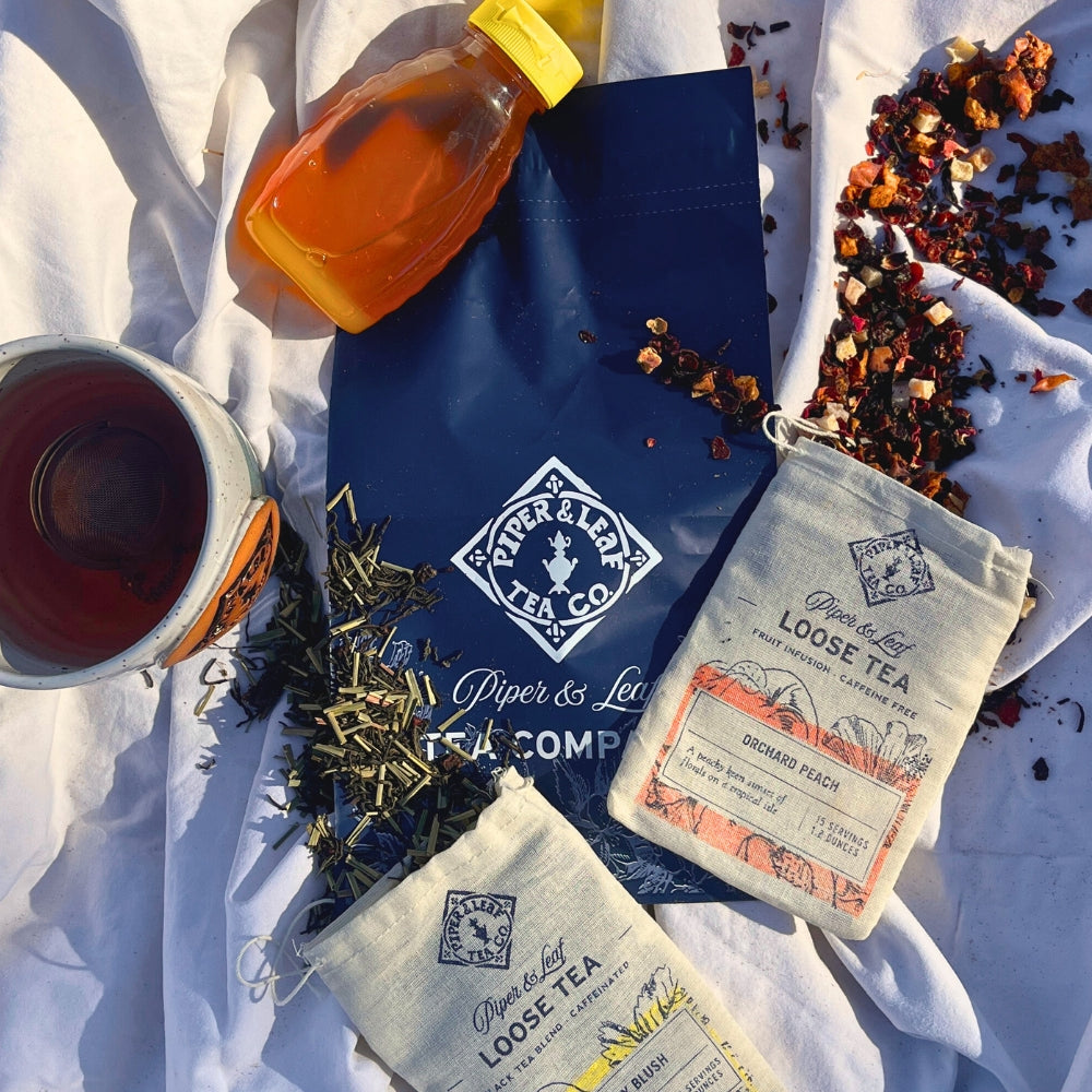 A Piper & Leaf Tea Co. Tea Subscription includes assorted loose tea bags, a cup of freshly brewed artisan tea, a jar of honey, and scattered tea leaves against a white background.