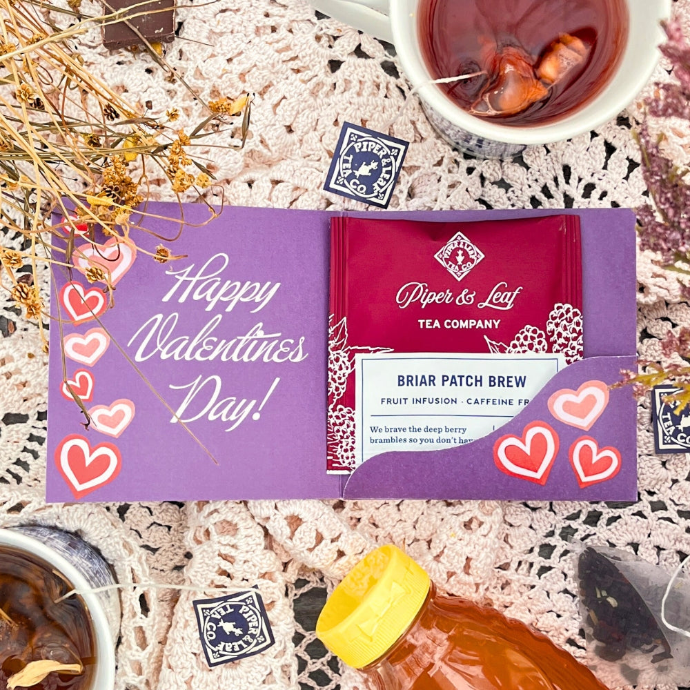 A romantic setup features Piper & Leaf Tea Co.'s Tea Valentines on a lace tablecloth with a "Happy Valentine's Day" card, heart stickers, and dried flowers, creating the perfect way to enjoy berry best blends.