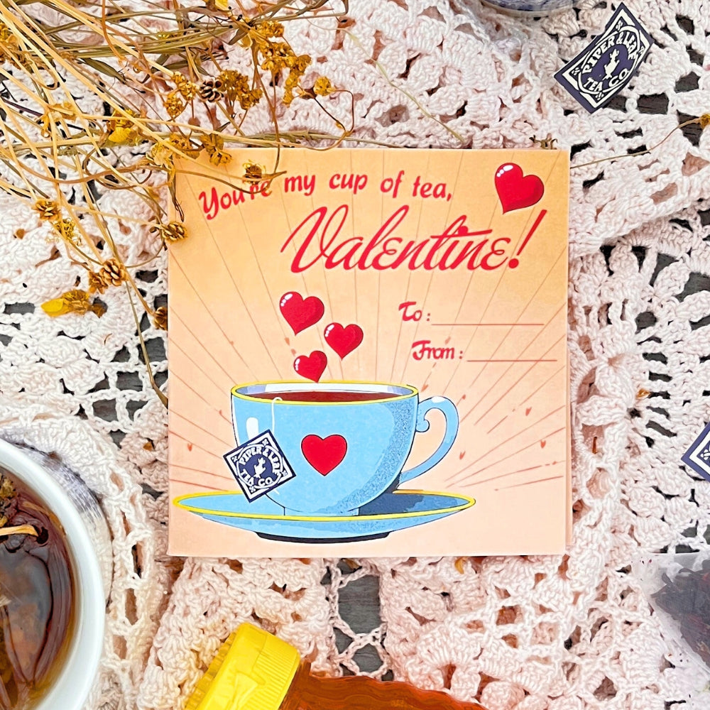 A Piper & Leaf Tea Co. "Tea Valentines" card features a teacup and hearts with the message, "You're my cup of tea, Valentine! To: From:" Displayed on lace with dried flowers and berry blends, this charming card captures the warmth of Valentine's Day tea gatherings.