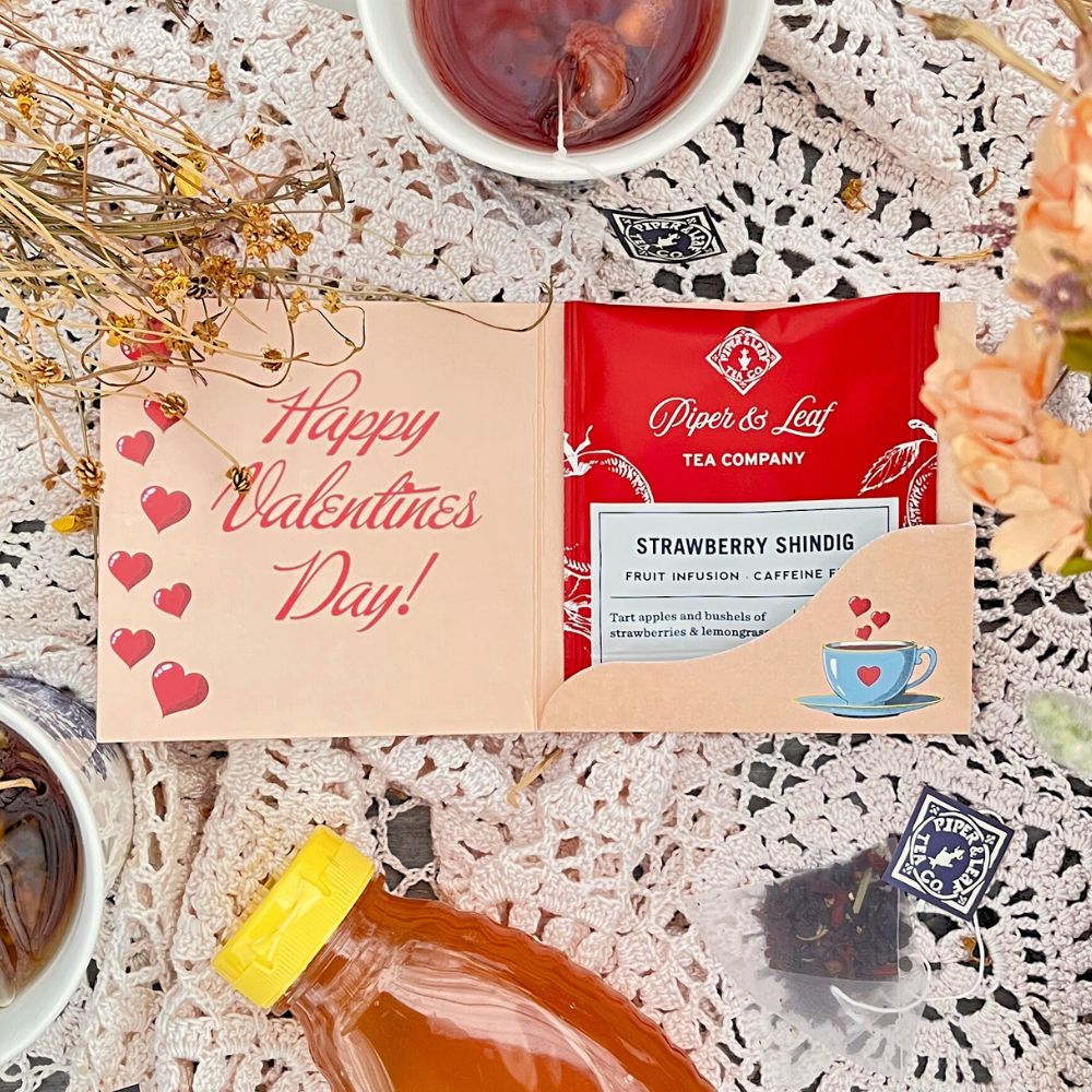 The Tea Valentines set by Piper & Leaf Tea Co. includes a "Happy Valentine's Day" card, a strawberry tea packet from our berry best blends, two tea cups, a honey bottle on a lace tablecloth, and decorative dried flowers for an enchanting touch.