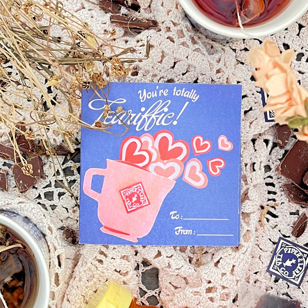 On a lace tablecloth, a card with a pink cup and heart design reads "You're totally terrific!" Surrounded by tea cups, chocolates, and dried flowers, it sets the scene for Tea Valentines by Piper & Leaf Tea Co., perfect for indulging in blends or creating your own tea bundle.