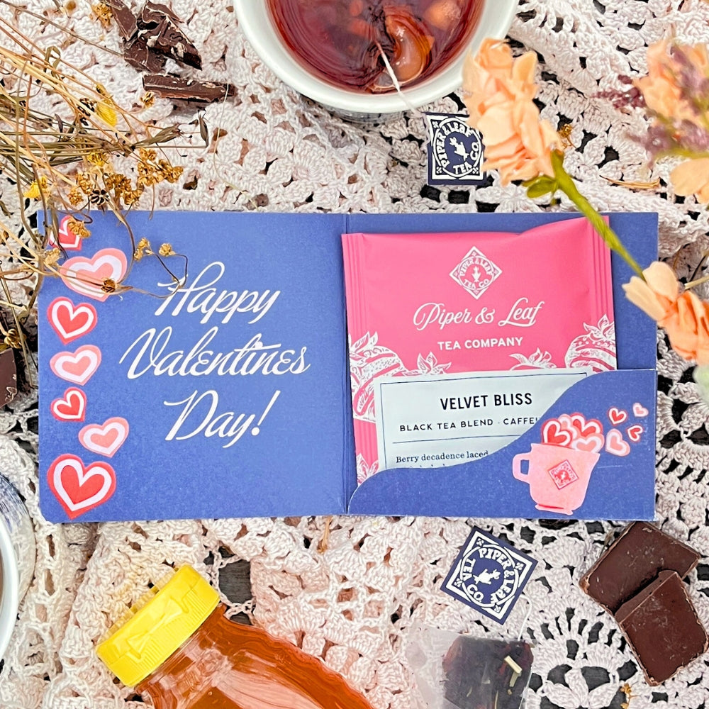 A Tea Valentines card by Piper & Leaf Tea Co. showcases a pink Velvet Bliss tea sachet on a lace tablecloth with hearts, chocolates, and flowers. Experience the love in every sip with our berry blends, ideal for creating your own personalized tea bundle.