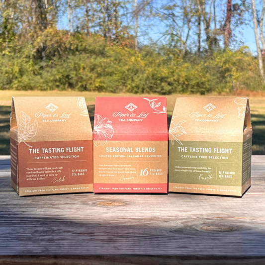 Three boxes from the Piper & Leaf Tea Co., known as the Tasting Flight Trio, are elegantly displayed outdoors on a wooden surface, inviting you to embark on a tea-riffic adventure.