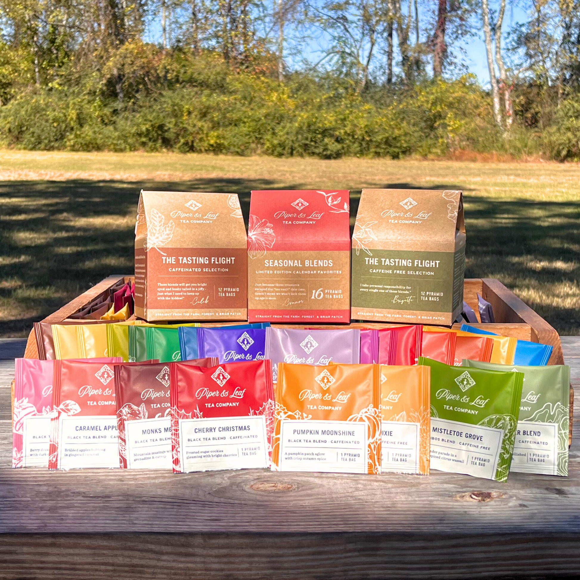 Embark on a delightful tea-tasting journey with Piper & Leaf Tea Co.'s Tasting Flight Trio. This charming collection of assorted tea packets and boxes is beautifully displayed on a wooden crate, nestled amidst trees and grass, beckoning you to savor the exquisite flavors in an outdoor setting.