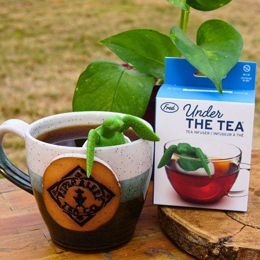 A green sea turtle-shaped tea infuser, branded as "Under the Tea Infuser" by Piper & Leaf Tea Co., floats on loose leaf tea. A labeled box in the background adds to the serene atmosphere with a plant and wooden surface completing your tranquil teatime experience.