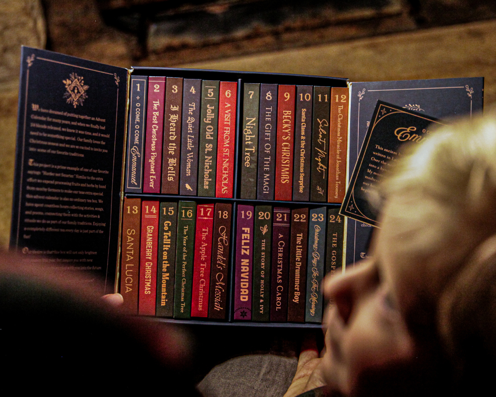 A person holds a festive book collection designed as an advent calendar with titles labeled for the 25 days of Christmas, such as "Santa Lucia" and "The Night Before Christmas." Beside the partially visible book cover, teas from the 24 Teas of Advent—Piper and Leaf Advent Calendar 2024 by Piper & Leaf Tea Co. are scattered around.