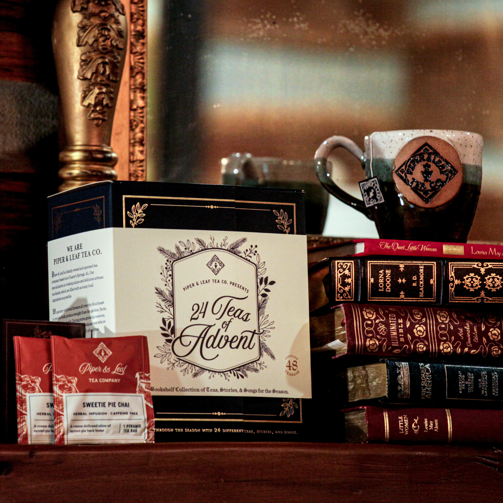 A display featuring a "24 Teas of Advent—Piper and Leaf Advent Calendar 2024" box set from Piper & Leaf Tea Co., various books with ornate covers, two packets of tea, and a mug placed in front of a mirror with a golden frame.