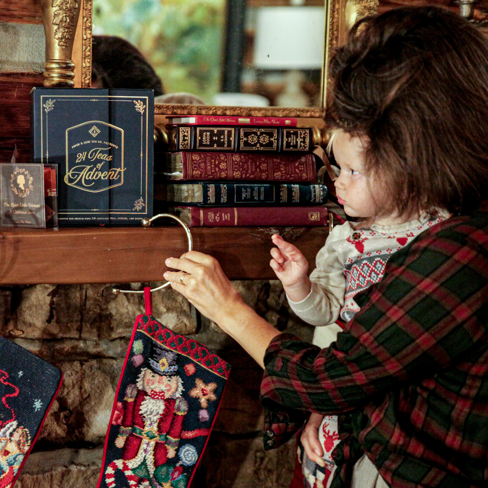 A child in festive clothing reaches for a Christmas stocking hanging from a mantle. The mantle, adorned with books and a framed photo, also features the Piper & Leaf Tea Co.'s 24 Teas of Advent—Piper and Leaf Advent Calendar 2024, adding to the holiday charm.