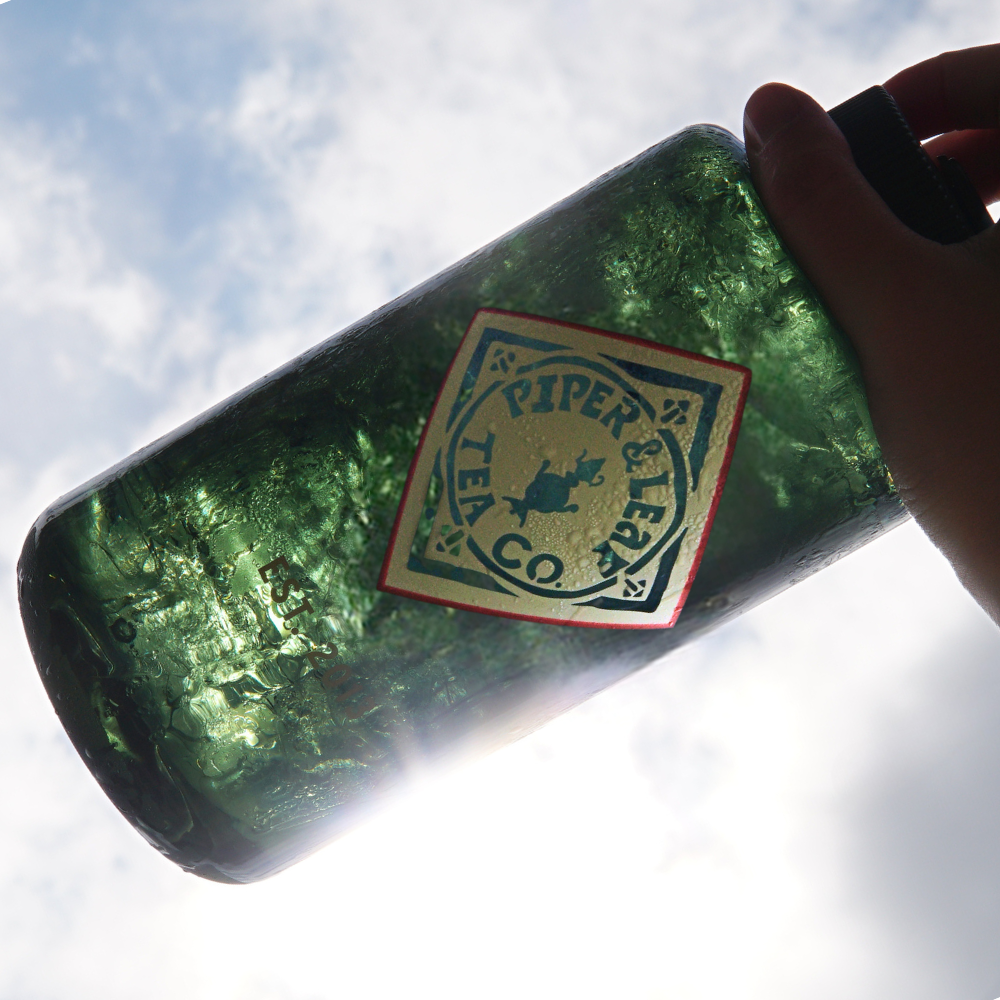 A hand holds a green, BPA-free 32oz Piper & Leaf Nalgene Water (Tea) Bottle - Retro Classic labeled "Piper & Leaf Tea Co." against a bright sky with clouds.