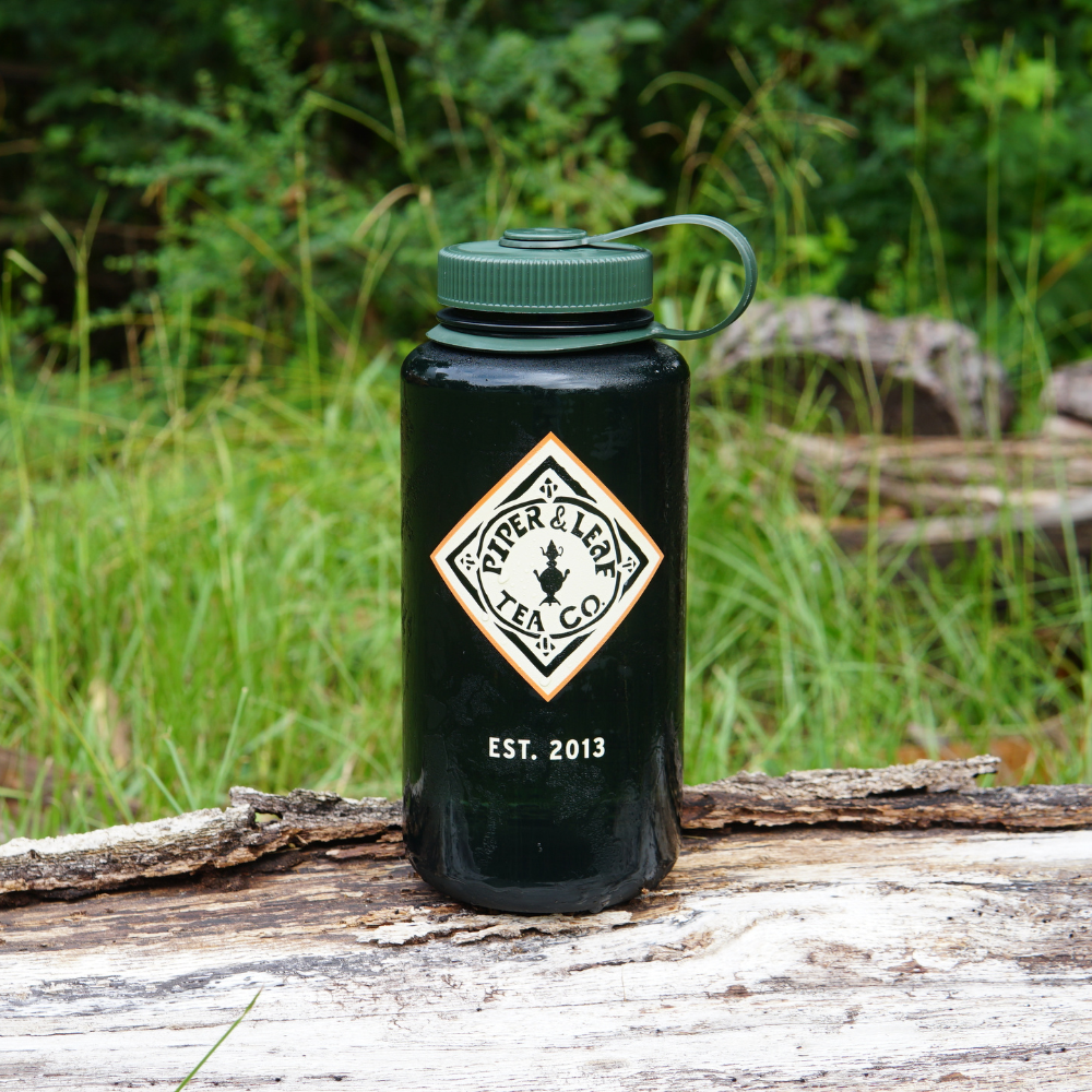 A 32oz Piper & Leaf Nalgene Water (Tea) Bottle - Retro Classic from Piper & Leaf Tea Co., featuring a handle and resting on a piece of wood in an outdoor setting. The text on the BPA Free, super durable bottle reads "EST. 2013.