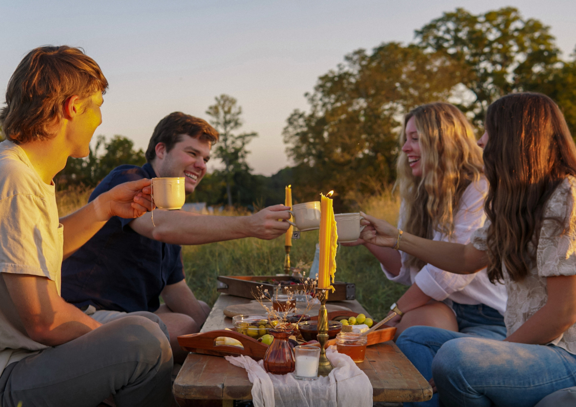 Four people toast with cups of Piper & Leaf Tea Co.'s Healthy Digestion blend at a low wooden table outdoors, surrounded by food and candles at sunset—an ideal way to enjoy digestive support together.