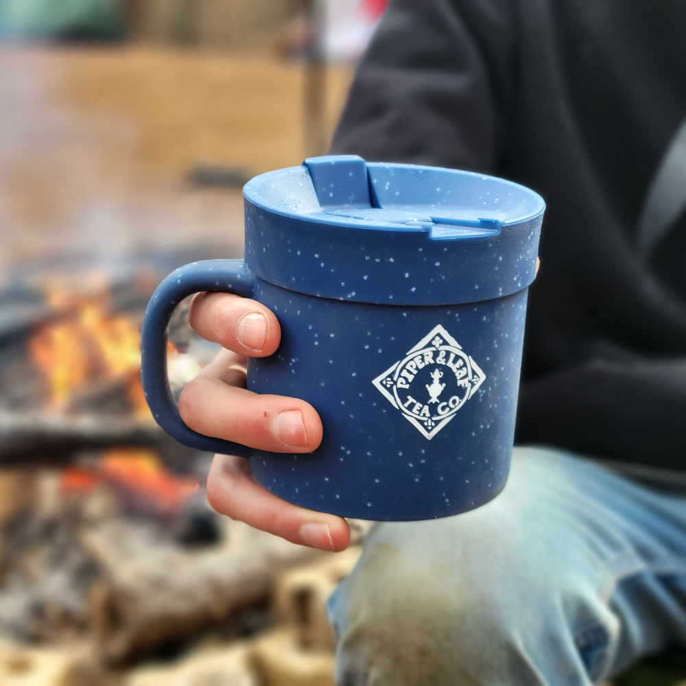 A person holding a blue enamel Limited Edition- P&L X Silipint 16oz Coffee Mug + Lid, featuring the Piper & Leaf Tea Co. logo, in front of a campfire.