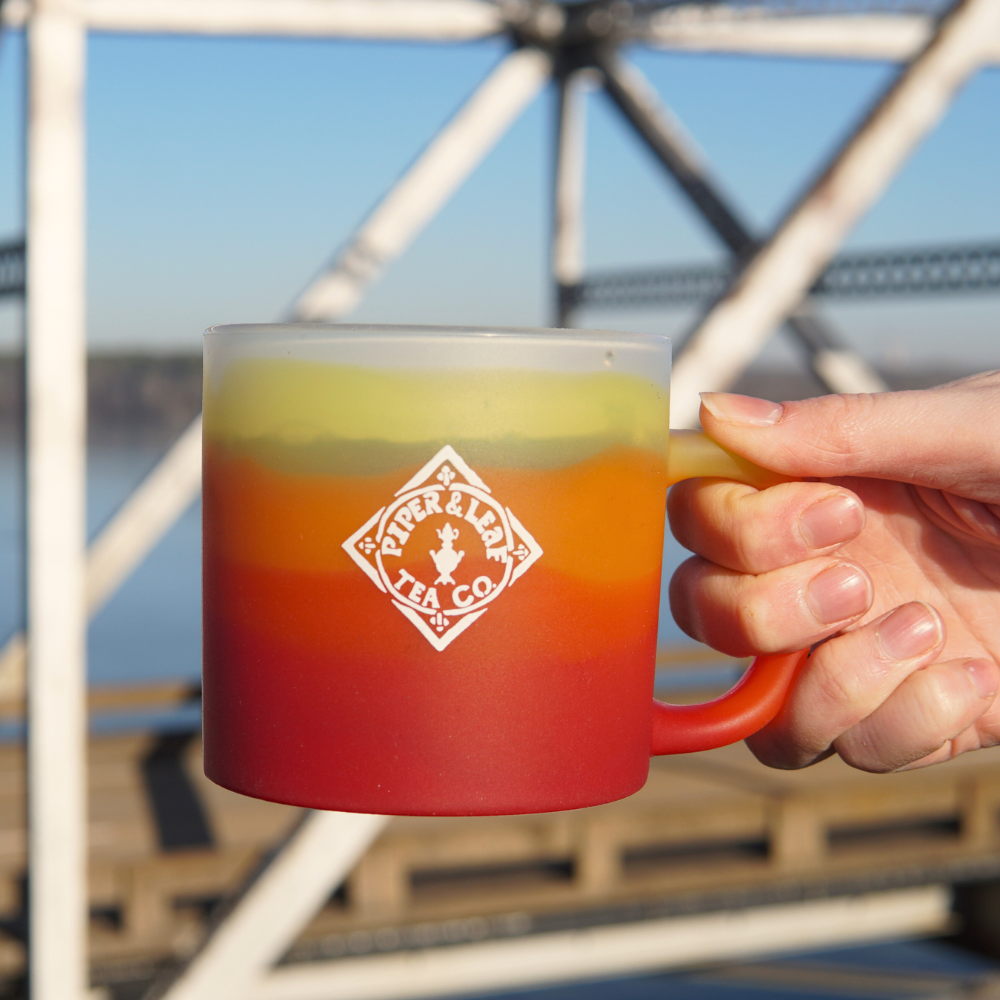 A hand holds a gradient red and yellow Piper & Leaf Tea Co. Limited Edition- P&L X Silipint 16oz Coffee Mug + Lid, crafted from BPA-free, platinum-cured silicone with the text "Piper & Leaf Tea Co." printed on it. In the background, a bridge and shimmering water create a dreamy, blurred effect.