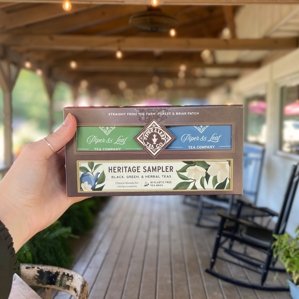 A hand holds a Piper and Leaf Tea Co. Heritage Sampler—Box of 18 Tea Bags—on a wooden porch with rocking chairs and greenery in the background.