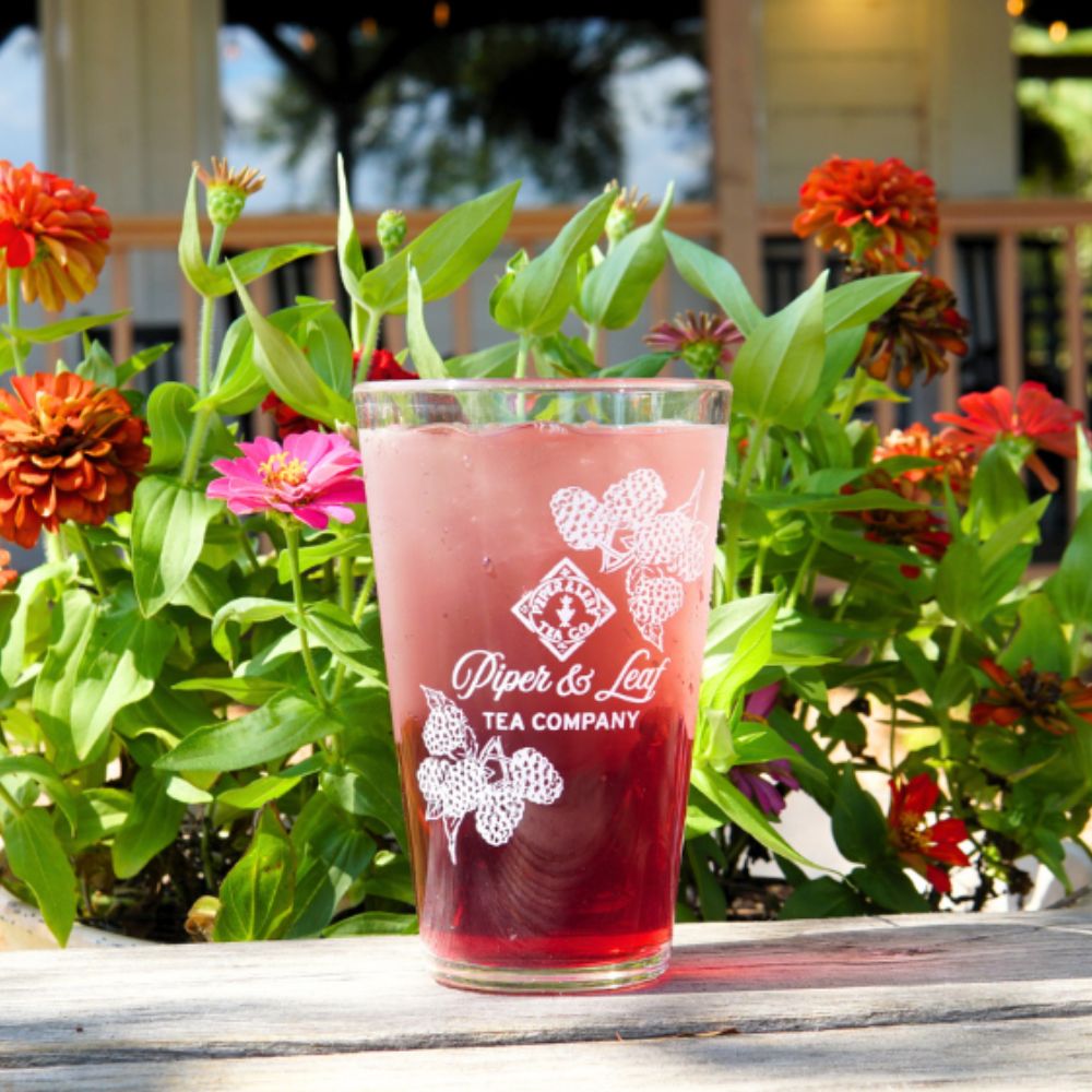 The Limited Edition: Into the Briar Patch P&L X Silipint 16oz Cup from Piper & Leaf Tea Co. elegantly holds a glass of red iced tea on a wooden table, surrounded by colorful flowers.