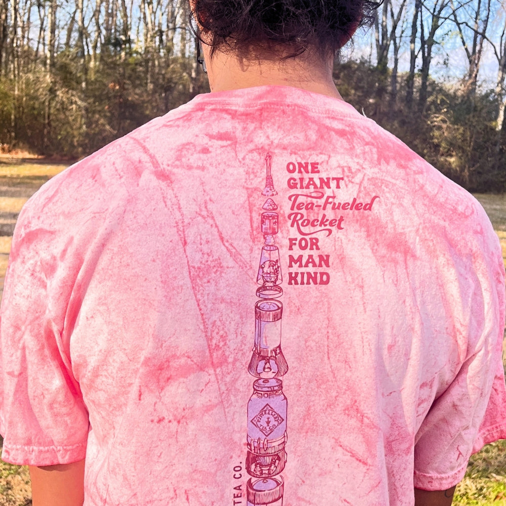 A person outdoors is wearing a Tie-Dye Retro Spaceman Tee by Piper & Leaf Tea Co., showcasing a pink tie-dye design with a rocket graphic and the text: "One giant tea-fueled rocket for mankind.