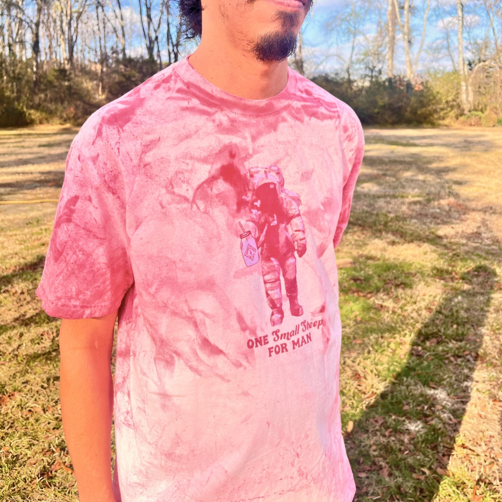 A person in a pink Piper & Leaf Tea Co. Tie-Dye Retro Spaceman Tee featuring an astronaut graphic and "One Small Step For Man" text stands outside amidst grass and softly rustling trees.