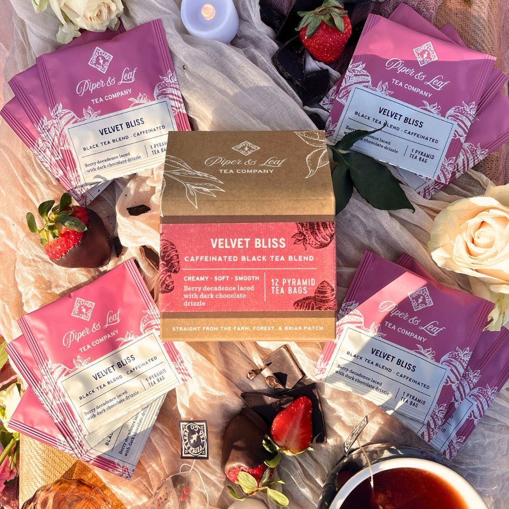 A box of 12 "Velvet Bliss" tea bags by Piper & Leaf Tea Co., Valentine's seasonal blend, is surrounded by roses, strawberries, and petals on a fabric background.