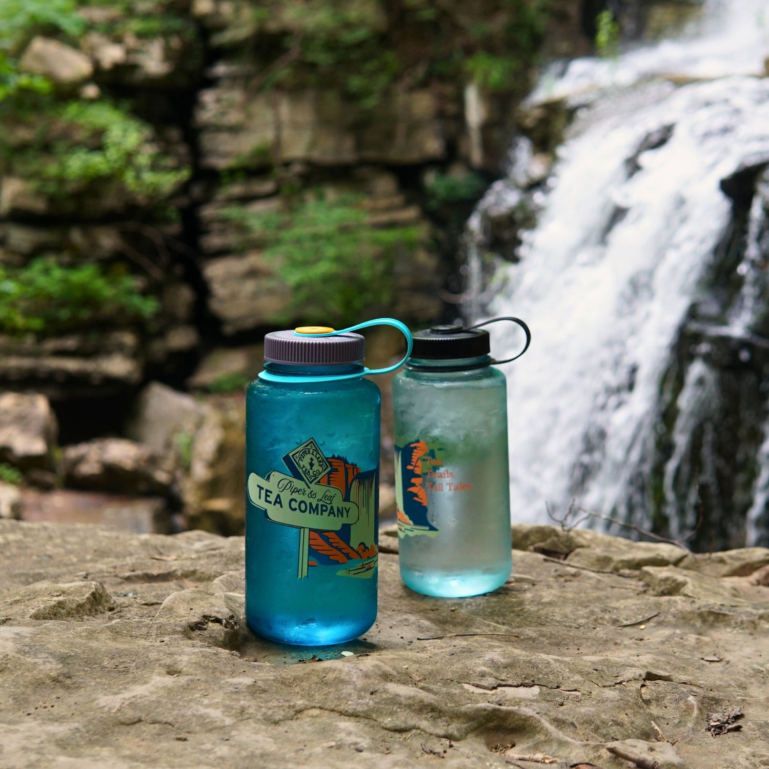 Two Piper & Leaf Tea Co. "Tea, Trails and Tall Tales" Nalgene bottles sit on a rocky ledge by a waterfall with lush greenery, ready for your next adventure.