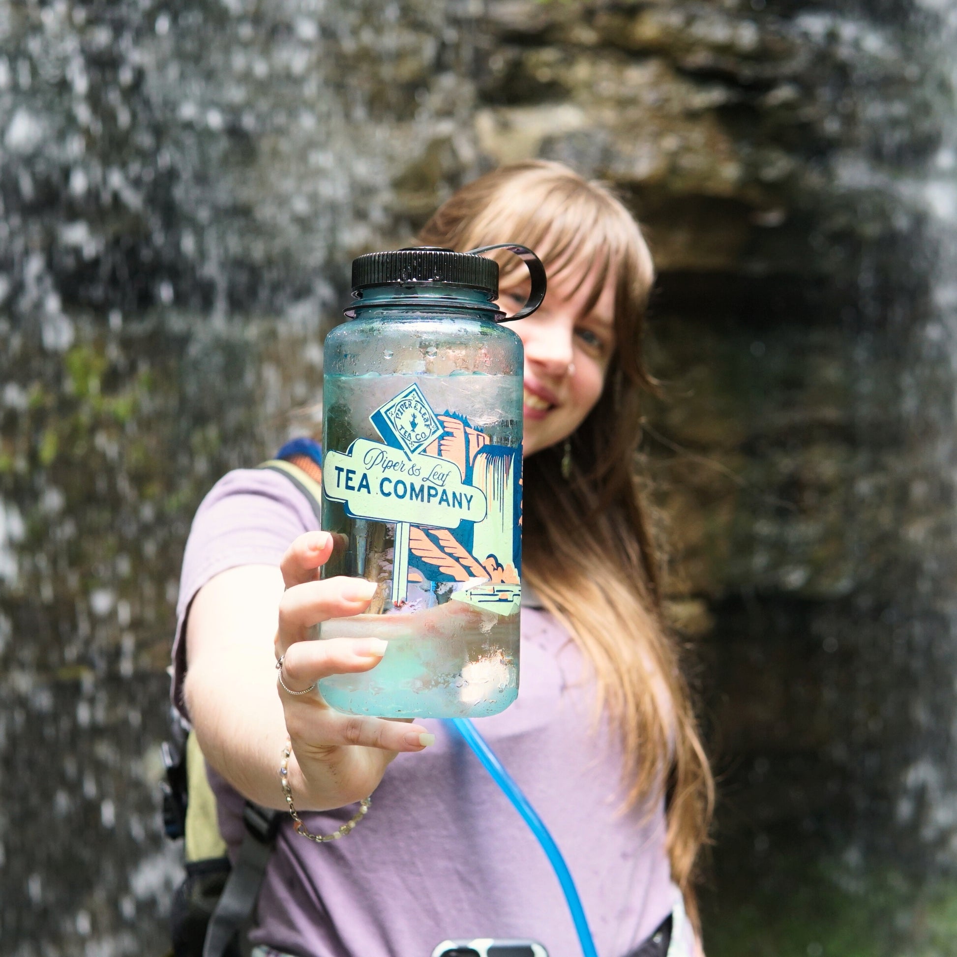 A woman with long hair stands by a waterfall, holding a clear "Tea, Trails and Tall Tales Nalgene" bottle from Piper & Leaf Tea Co. toward the camera, capturing an adventurous outdoor spirit.