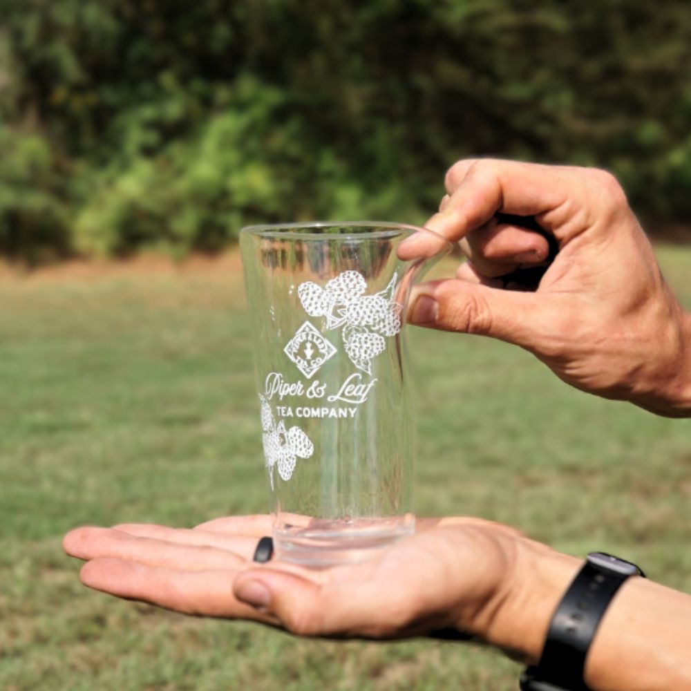 Two hands holding a Limited Edition: Into the Briar Patch P&L X Silipint 16oz Cup from Piper & Leaf Tea Co., with trees and grass in the background.