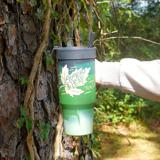 Against a vine-covered tree, someone holds a Piper & Leaf Tea Co. Limited Edition - Autumn Leaf P&L X Silipint 32oz Tumbler with Handle, ideal for seasonal sipping.