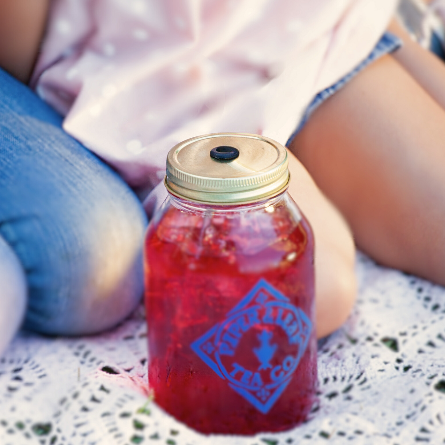 A girl sitting on a blanket next to a Mason jar filled with liquid and a Piper & Leaf Tea Co. Drinking Lid for Mason Jar.