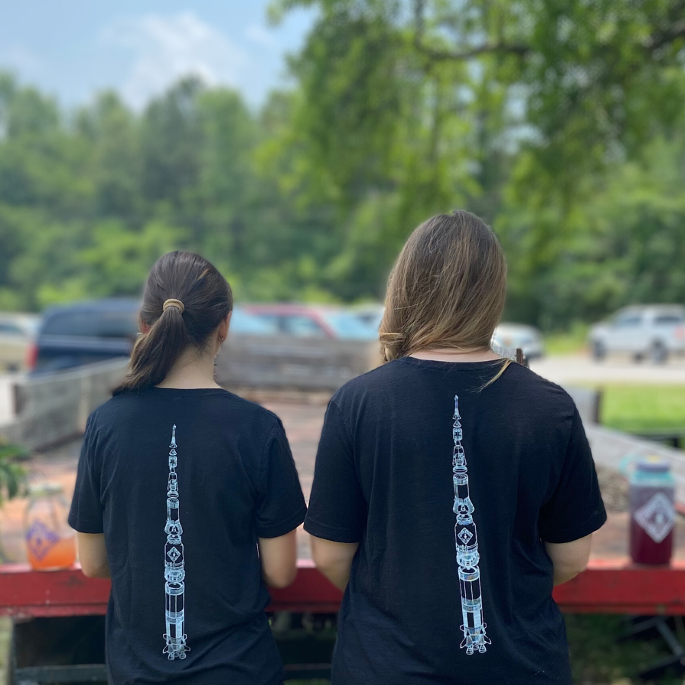 Two individuals with their backs to the camera, wearing matching Piper & Leaf Tea Co. Limited Edition 3D Vision Space Man Tees with a white tower graphic, standing outdoors.