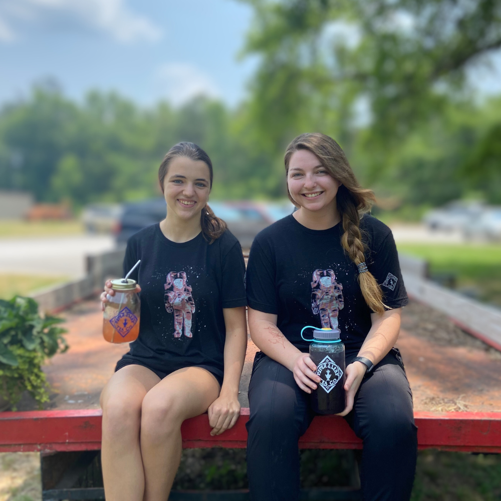Two individuals sitting on a bench holding drinks, wearing black t-shirts with a Piper & Leaf Tea Co. Limited Edition 3D Vision Space Man Tee graphic print, and smiling at the camera.