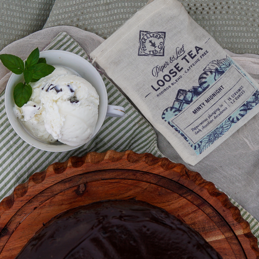A cup of vanilla ice cream with mint leaves sits beside a Piper & Leaf Tea Co. Minty Midnight Muslin Bag of Loose Leaf Tea (15 Servings) and a rich chocolate cake on a wooden tray.