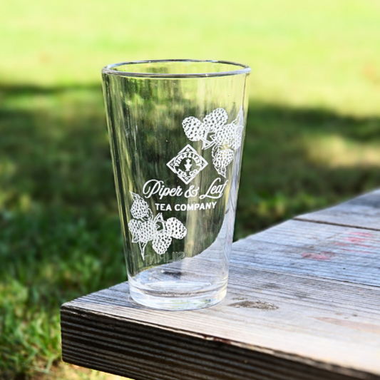 A Limited Edition: Into the Briar Patch P&L X Silipint 16oz Cup from Piper & Leaf Tea Co., featuring a floral design and logo, rests on a wooden bench outside, highlighting its unbreakable drinkware quality against the grassy backdrop.