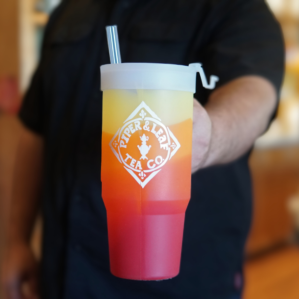 A person holding a colorful iced tea in a clear, Piper & Leaf Tea Co. limited edition P&L X Silipint 32oz Tumbler with Handle with a metal straw.