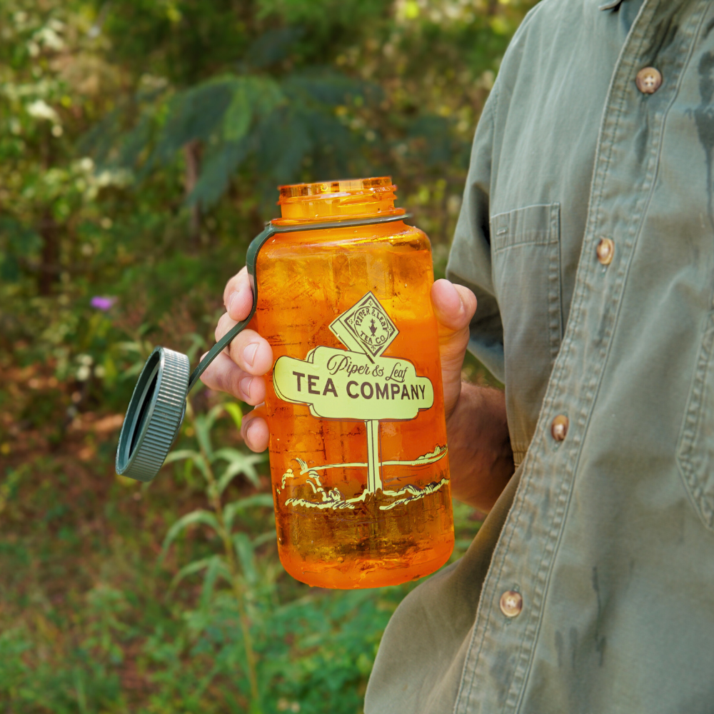 A person in a green shirt holds an orange, BPA-free Piper & Leaf Nalgene Water (Tea) Bottle: Road Sign with a gray lid in an outdoor setting, showcasing one of the super durable Fall Nalgenes from Piper & Leaf Tea Co.