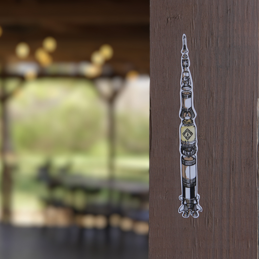 An ornate metal mezuzah affixed to a wooden doorpost, adorned with a "P&L Saturn V Rocket Fuel 6" Sticker Two Pack" sticker, with a blurred background.