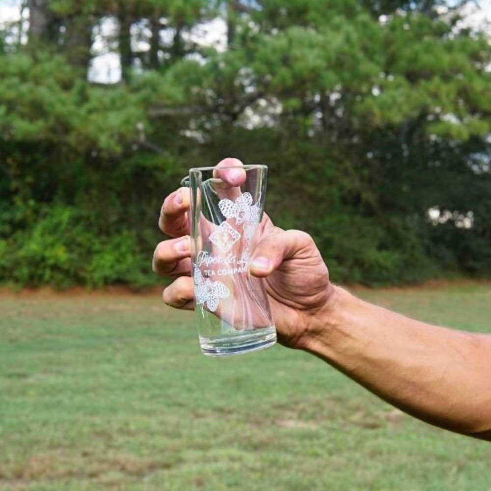 A hand holds a Limited Edition: Into the Briar Patch P&L X Silipint 16oz Cup from Piper & Leaf Tea Co., showcasing its decorative design as unbreakable drinkware ideal for outdoor settings, with a grassy area and trees in the background.