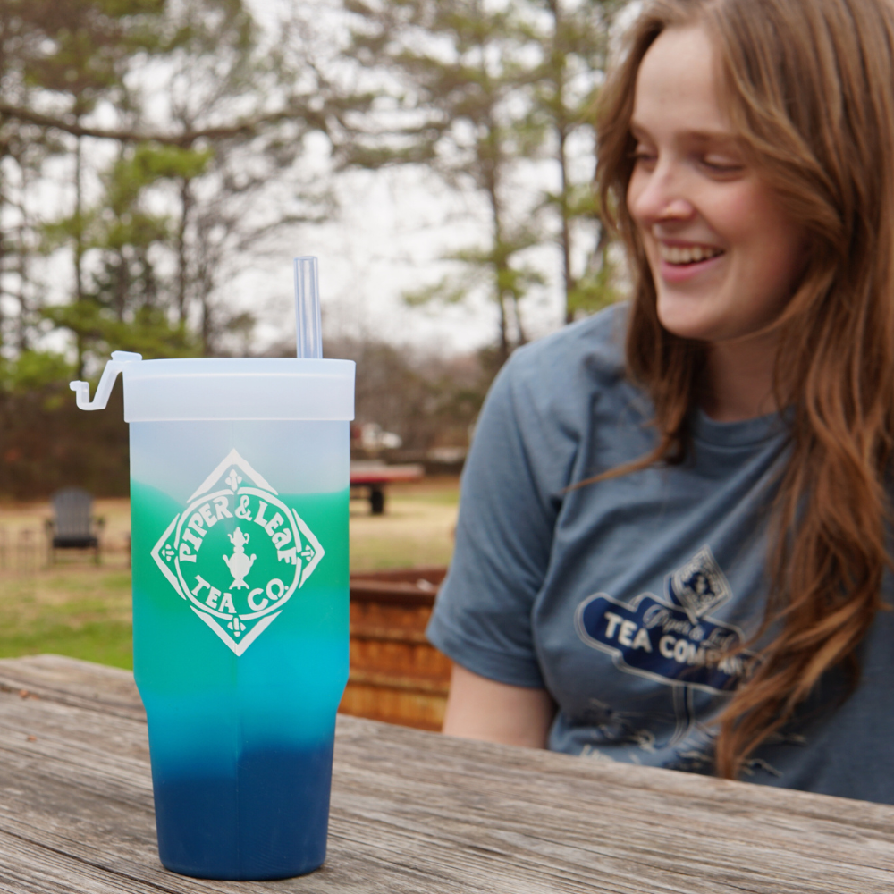 A young woman smiling and looking at a Limited Edition- P&L X Silipint 32oz Tumbler with Handle in blue gradient from Piper & Leaf Tea Co. on a wooden surface outdoors.