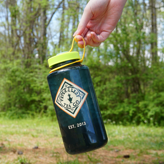 A hand holding a blue BPA free Piper & Leaf Nalgene water bottle with a "Piper & Leaf Tea Co." label outdoors.