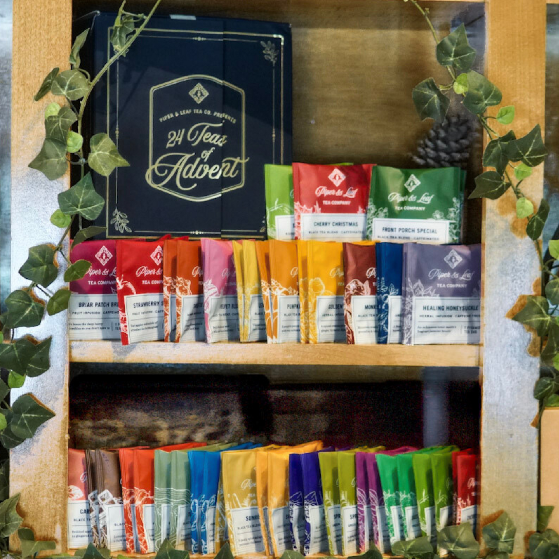 A wooden shelf displays a Piper & Leaf Tea Co. Advent Calendar Refill Kit and rows of colorful artisan tea bags, all surrounded by green ivy decor.