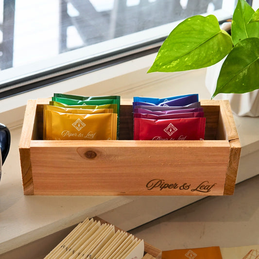 A "Wooden Steep Box" by Piper & Leaf Tea Co. holds colorful tea packets on a sunlit windowsill, with a vibrant green plant adding freshness on the right side.