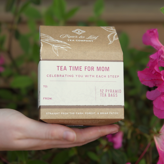 A person holding a box of "Tea Time For Mom- Box of 12 Individually Wrapped Tea Bags" from Piper & Leaf Tea Co., surrounded by pink flowers.