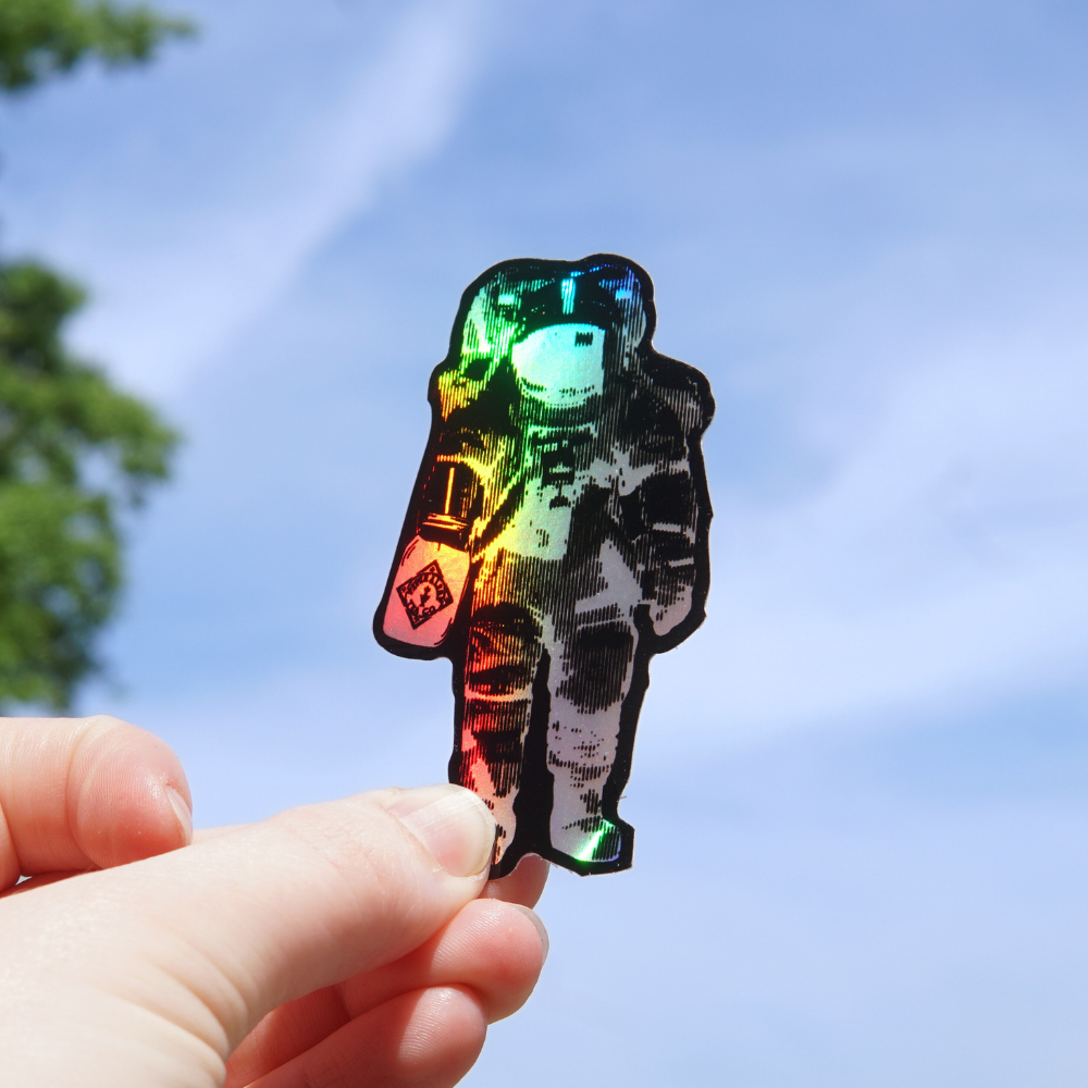 A hand holding a Piper and Leaf Tea Co. P&L Spaceman Holographic Sticker against a clear blue sky.