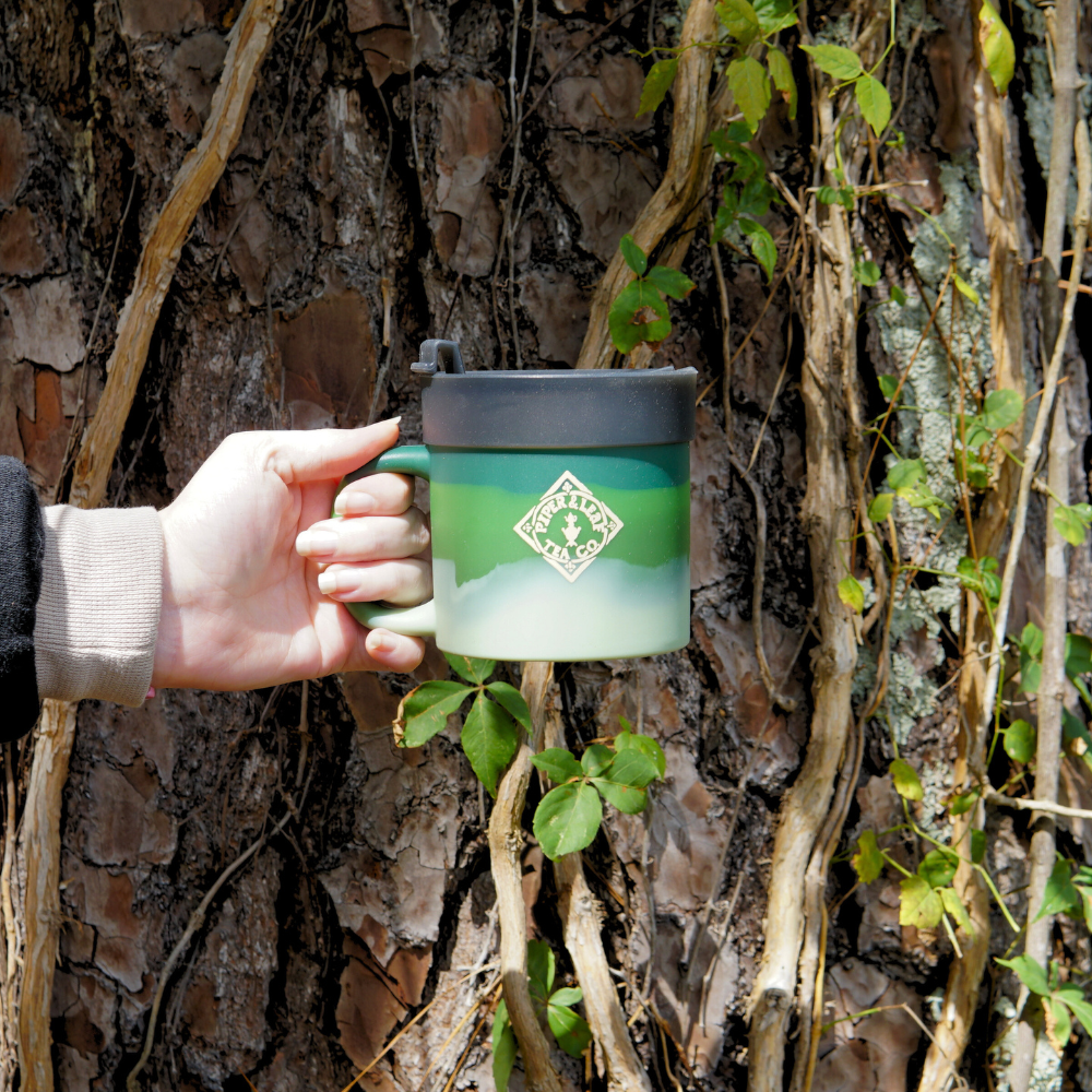 A hand holding the Limited Edition: Autumn P&L X Silipint 16oz Coffee Mug + Lid by Piper & Leaf Tea Co., set against a textured tree bark backdrop entwined with vines.