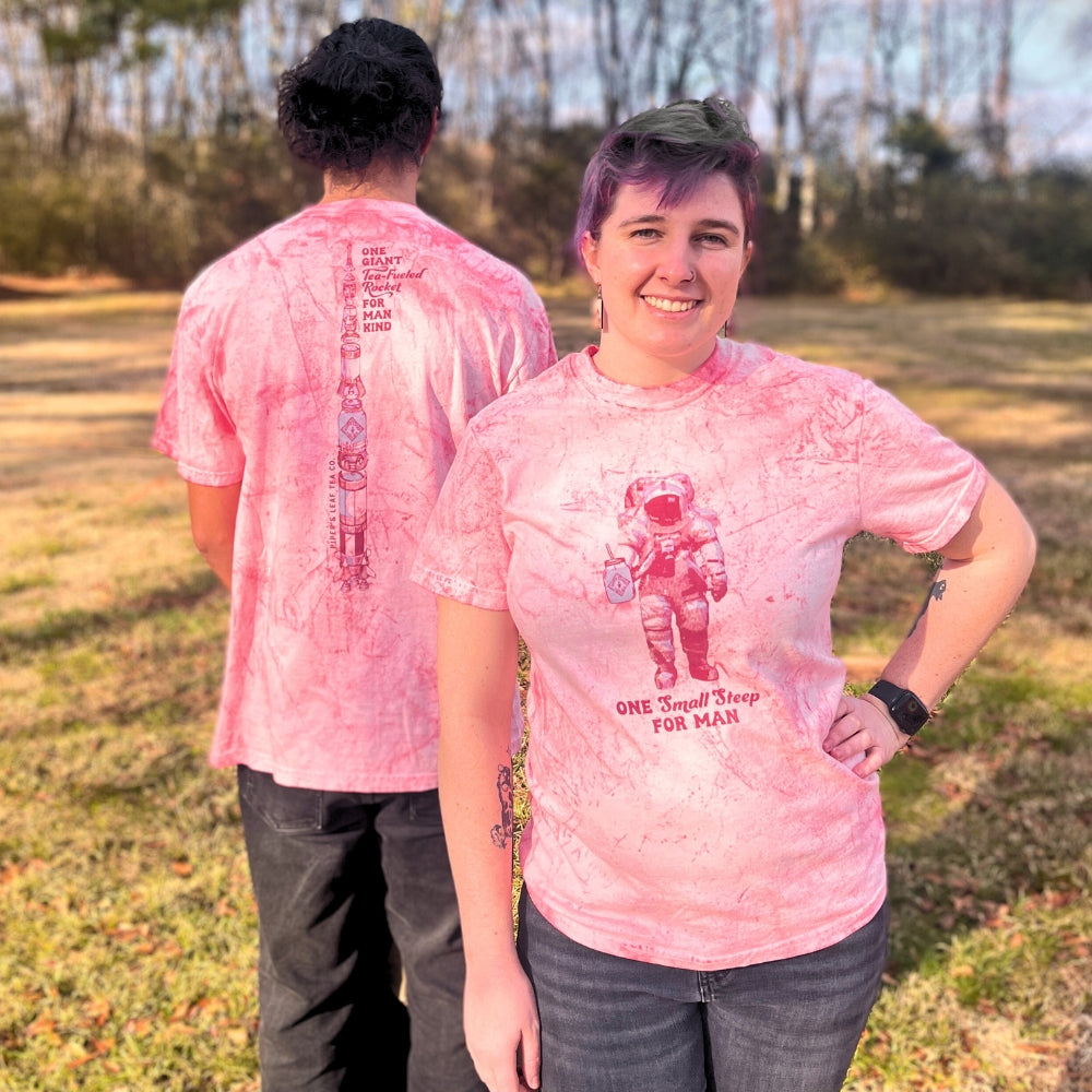 Two people wearing Tie-Dye Retro Spaceman Tees from Piper & Leaf Tea Co. stand outdoors, with one facing forward and the other showing their back to the camera, exuding a cool cosmic vibe.