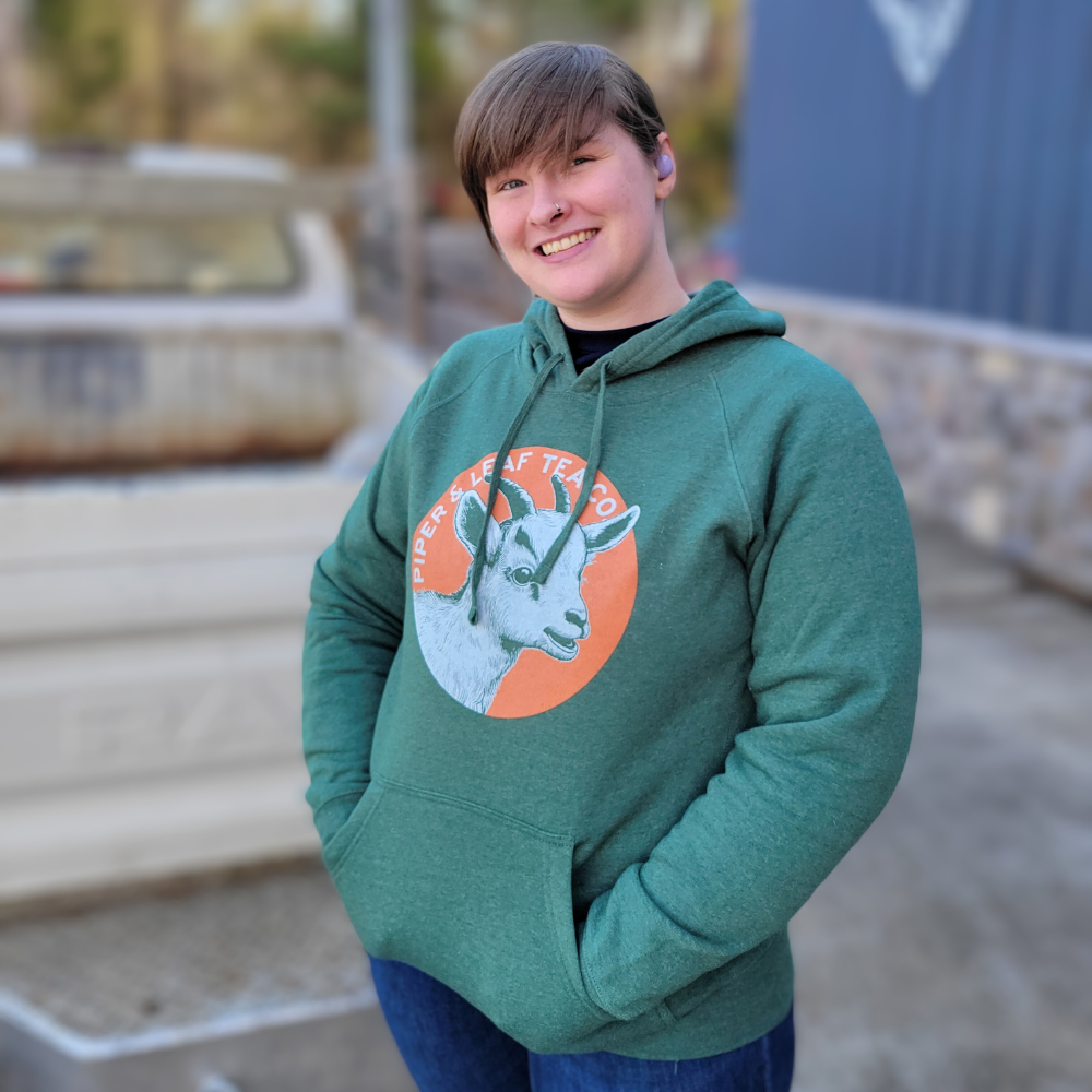 An adult-sized woman wearing a green Goat Hoodie by Piper & Leaf Tea Co. with a Clyde-inspired goat on it.