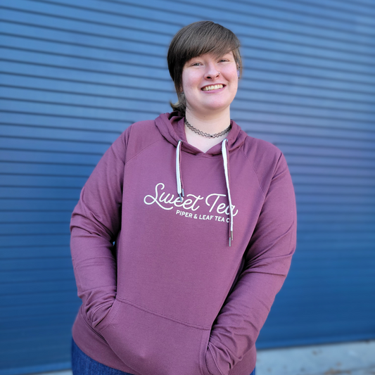 A woman wearing a Piper & Leaf Tea Co. Sweet Tea Lightweight Hoodie smiles in front of a blue wall.