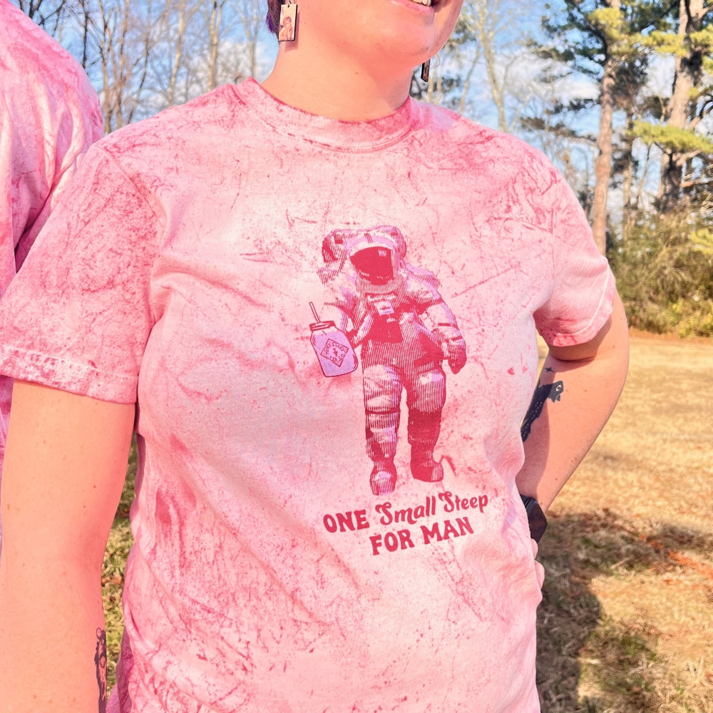 A person wearing a Piper & Leaf Tea Co. "Tie-Dye Retro Spaceman Tee" featuring an astronaut graphic and the text "One Small Steep for Man" is standing outside.