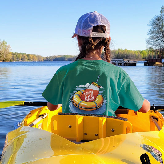 Wearing a Piper & Leaf Tea Co. Life Preserver T-Shirt and a cap, someone with braided hair paddles a yellow kayak on a calm, tree-lined lake, savoring a peaceful summer adventure.