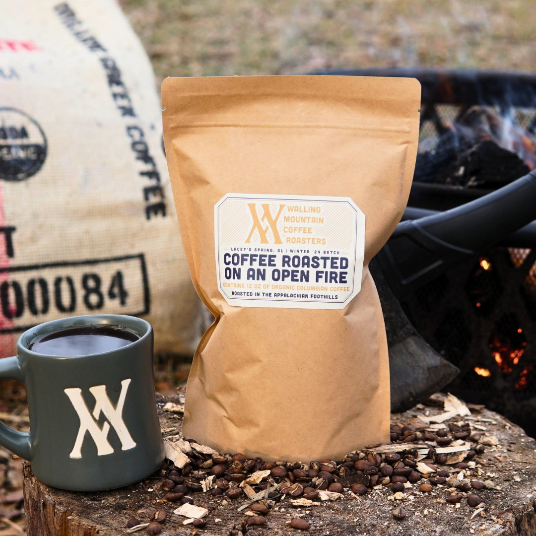 A 12oz Bag of Campfire Roast Coffee from Walling Mtn. sits beside a mug and scattered beans, with a burlap sack and outdoor setting in the background. Savor the rich flavors of our organic Colombian/Peruvian blend crafted using traditional roasting methods.