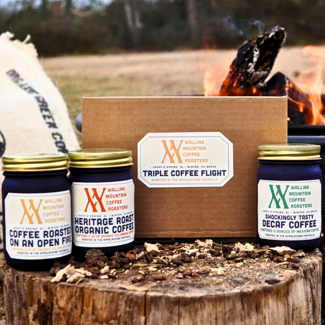 A coffee set featuring two jars labeled "Walling Mountain Farms Heritage Roast Organic Coffee" and "Shockingly Tasty Decaf," alongside a box titled "Triple Coffee Flight — Walling Mtn." by Walling Mtn. Coffee, is shown outdoors near a fire—ideal for fans of an open-fire roast experience.