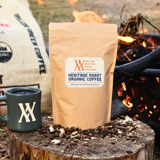 A brown 12oz bag of Walling Mtn. Coffee's Heritage Roast rests on a tree stump beside a logo-bearing green mug, amidst scattered beans and a cozy fire in the background, creating an inviting scene.